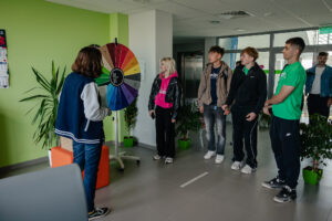 Korytarz. Po lewej kolorowe koło fortuny i stojąca tyłem do kadru osoba z obsługi. Po prawej (przodem do kadru) czworo uczestników gry „Naukowy Detektyw – Misja w Terenie”. W tle przeszklone drzwi wejściowe i kwiaty doniczkowe.