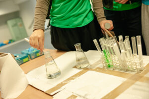 Na stole probówki i kolby. Jedna z uczestniczek (ujęcie na same ręce) wlewa zawartość probówki do kolby, wypełniając zadanie w grze „Naukowy Detektyw – Misja w Terenie”.
