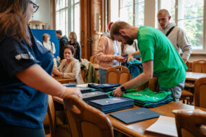 Ujęcie bokiem. Na drewnianym stole granatowe, niebieskie i zielone koszulki z napisem: explorUS! Nauka bez granic oraz grantowe teczki z białym logiem 40-lecia Uniwersytetu Szczecińskiego. Dookoła stojący ludzie. W tle okna.