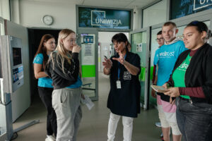 Kilkoro uczniów w niebieskich i zielonych koszulkach z napisem: „explorUS! Nauka bez granic” stoi na korytarzu i słucha, na czym polega praca w uniwersyteckim radiu i telewizji. W środku w ciemnogranatowej koszulce z logiem UNIwizji szefowa jednostek. W tle drzwi, nad którymi umieszczono logo UNIwizji.