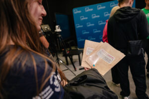 Ujęcie bokiem. Dziewczyna po lewej stronie trzyma beżowe teczki stylizowane na postarzone. Na teczkach napis: Dziennik badaczy, Naukowy Detektyw – Misja w Terenie. W tle od lewej: kamera na statywie, granatowa ścianka z białymi logami UNIwizji – Telewizji Uniwersytetu Szczecińskiego i stojący ludzie.