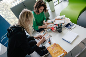 Ujęcie z góry. Dwie kobiety (jedna przodem do kadru, druga bokiem do kadru) siedzą przy białym stole i wpisują informacje w Dziennikach badawczych gry Naukowy Detektyw – Misja w Terenie. Na stole dzienniki, ulotki, długopisy w kubku, notes i pieczątka.