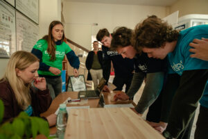 Korytarz. Pośrodku podłużny stół. Po lewej dwie studentki koordynujące zadanie. Po prawej pochyleni nad stołem trzej uczniowie w granatowych i niebieskich koszulkach z napisem: „explorUS! Nauka bez granic”, którzy wypełniają zadanie w grze „Naukowy Detektyw – Misja w Terenie”.