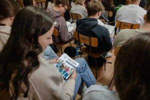 Ujęcie tyłem na uczniów siedzących na drewnianych numerowanych krzesłach. Uczniowie przeglądają informatory Uniwersytetu Szczecińskiego dla kandydatów na studia.