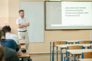 Klasa szkolna. Na zdjęciu prelegent. Za nim biała pionowa tablica. Po prawej ekran z prezentacją. Na pierwszym planie po lewej uczniowie w ławkach (ujęcie tyłem), po prawej puste ławki numerowanymi drewnianymi krzesłami.