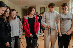 Korytarz szkolny. Pięcioro uczniów (3 uczennice po lewej i 2 uczniów po prawej) z nauczycielką (w środku) robią zadanie, trzymając długie sznurki.