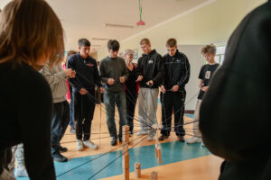 Korytarz szkolny. Sześcioro uczniów z nauczycielką (w środku) robi zadanie, trzymając długie sznurki, do których na środku przyczepiony jest jeden ciężarek.