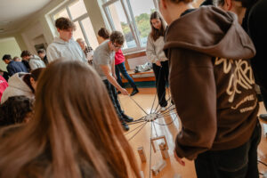 Korytarz szkolny. Uczniowie robią zadanie, trzymając długie sznurki, do których na środku przyczepiony jest jeden ciężarek.