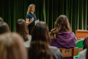 Ujęcie tyłem na uczniów siedzących na drewnianych numerowanych krzesłach. W tle przedstawicielka Uniwersytetu Szczecińskiego prezentująca ofertę dla kandydatów na studia. Za nią ciemnozielona kurtyna.