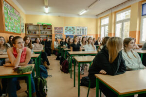 Klasa szkolna (ujęcie przodem). Od lewej uczniowie w ławkach, po prawej okna. W tle szkolne plansze edukacyjne i szafki.