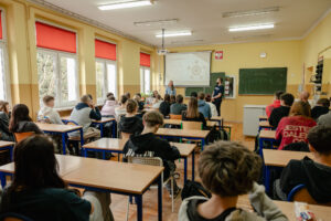 Klasa szkolna. Po lewej okna, dalej uczniowie w ławkach. W tle zielone tablice, ekran z prezentacją i troje prelegentów.