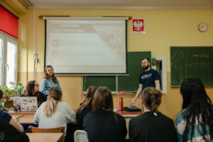 Klasa szkolna. Na pierwszym planie kilkoro uczniów tyłem do kadru. Po lewej okno. Dalej troje prelegentów i zielona tablica. U góry ekran z prezentacją.