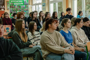 Klasa szkolna (ujęcie bokiem). Od lewej uczniowie w ławkach, po prawej okna. W tle szkolne plansze edukacyjne.