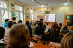 Klasa szkolna. Po lewej okna, dalej uczniowie w ławkach (tyłem do kadru). W tle ekran z prezentacją i dwoje prelegentów (kobieta po prawej i mężczyzna po lewej).