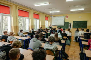 Klasa szkolna. Po lewej okna, dalej uczniowie w ławkach (tyłem do kadru). W tle zielone tablice, ekran z prezentacją i prelegentka.