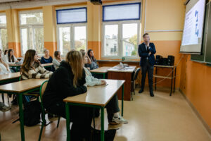 Klasa szkolna. Na pierwszym planie uczniowie w ławkach (ujęcie bokiem). Po prawej prelegent patrzący na ekran z prezentacją. W tle okna.