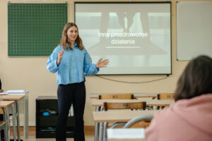 Klasa szkolna. Na zdjęciu prelegentka. Za nią zielona tablica w kratkę po lewej i po prawej ekran z napisem: „Inne prozdrowotne działania”. Obok niej ławki z numerowanymi drewnianymi krzesłami.