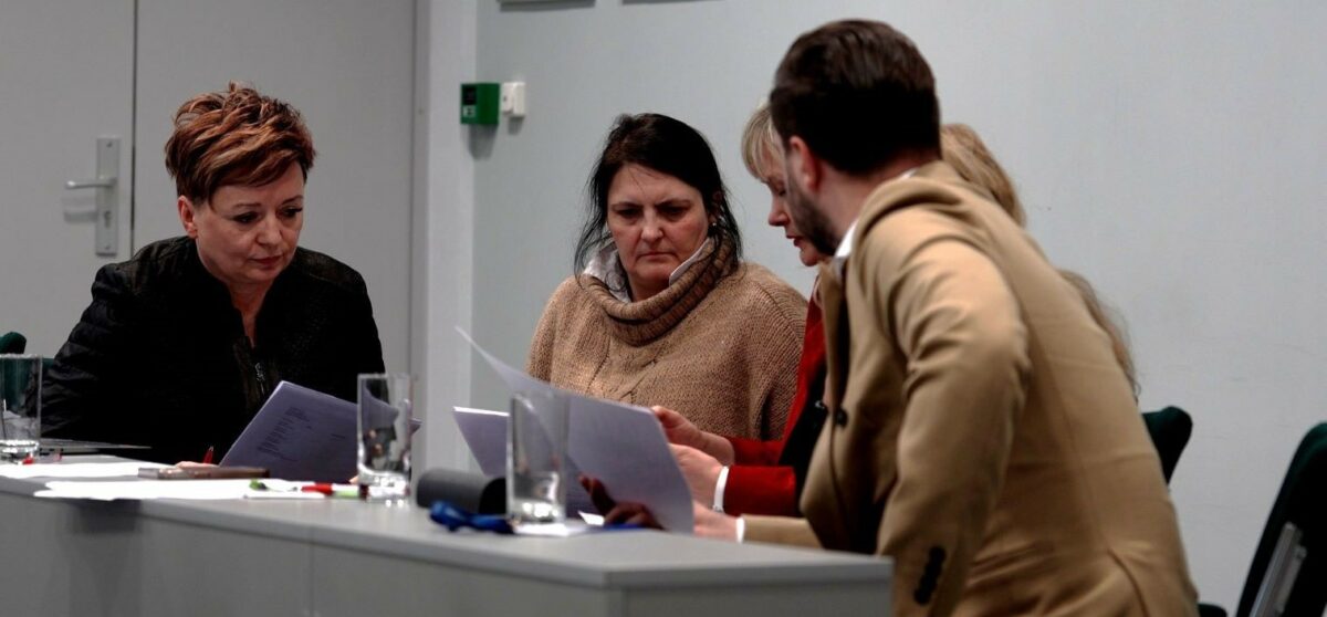 Grupa czterech osób siedzących przy stole konferencyjnym w pomieszczeniu biurowym. Wszyscy mają skupione, poważne miny i trzymają kartki papieru, jak podczas spotkania lub narady. Po lewej stronie siedzi kobieta o krótkich, rudobrązowych włosach w ciemnej marynarce, pochylona nad dokumentem. Na środku siedzi kobieta o ciemnych włosach w beżowym, grubym swetrze z wysokim kołnierzem, patrząca na kartki. Obok niej siedzi kobieta w czerwonym ubraniu, częściowo zasłonięta, także czytająca dokument. Po prawej stronie, na pierwszym planie, siedzi mężczyzna w beżowej marynarce, odwrócony bokiem do reszty. Na stole stoją przezroczyste szklanki z wodą oraz leżą długopisy i papiery. W tle widać jasnoszarą ścianę i drzwi; brak czytelnych napisów.