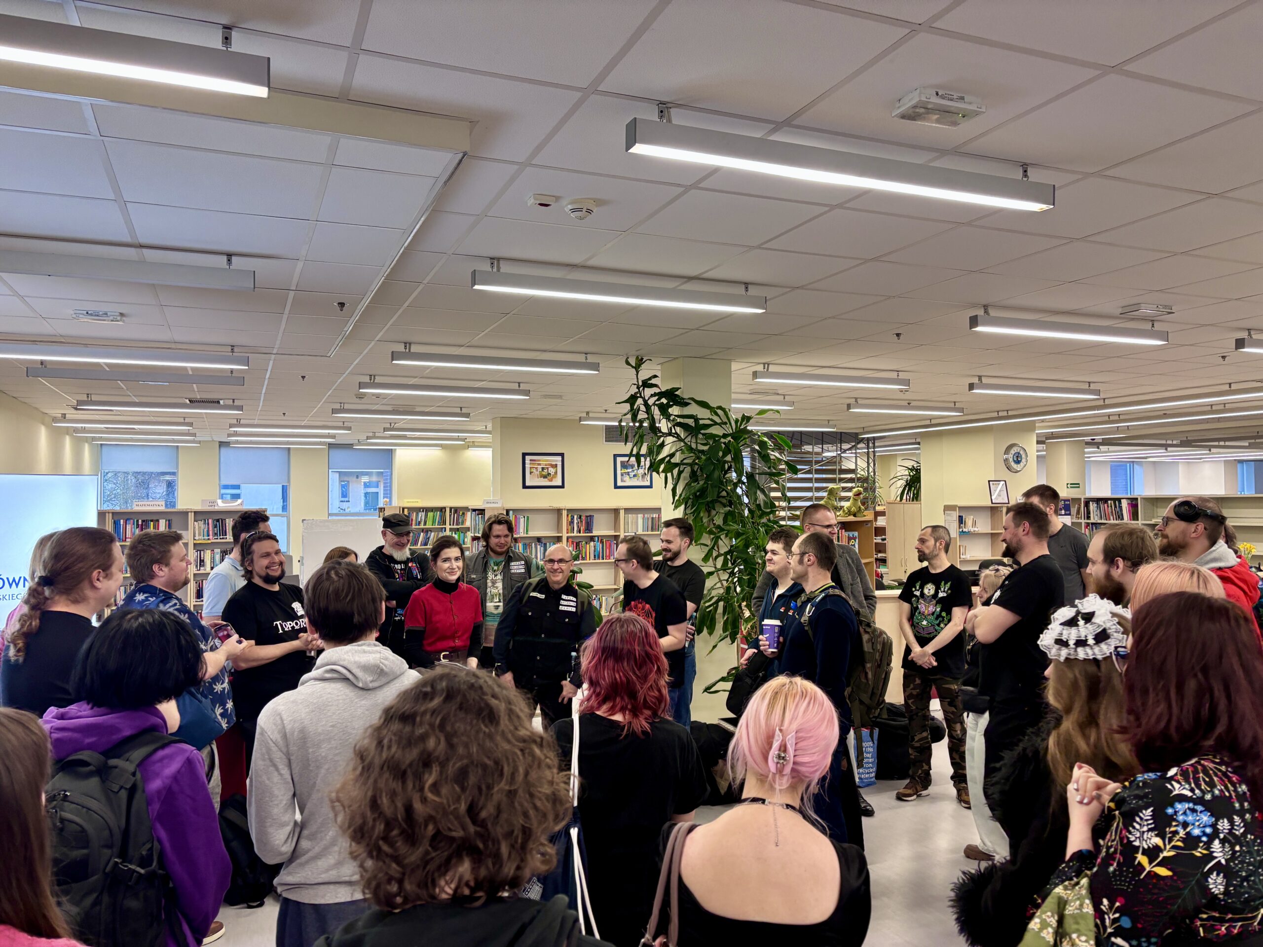 Grupa ludzi stojących w dużej sali biblioteki, słuchających mężczyzny w centrum.