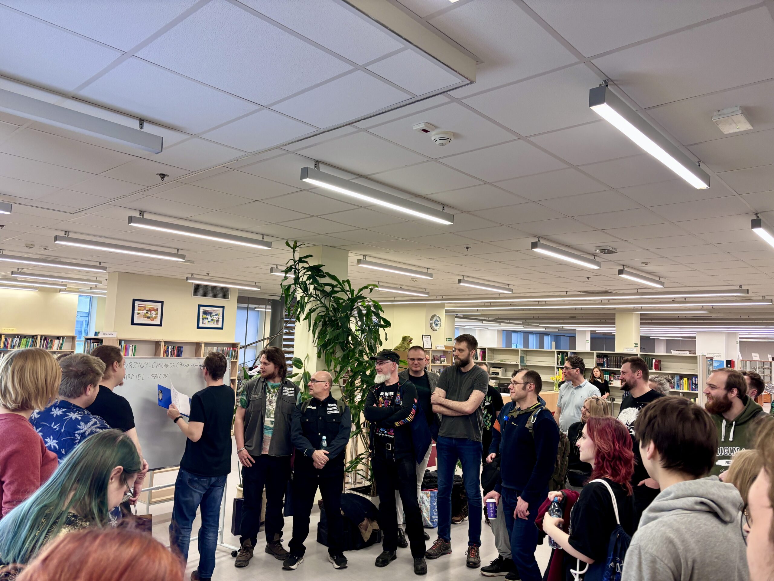 Grupa osób w sali biblioteki słuchająca mężczyzny, który wyjaśnia coś posiłkując się tablicą.