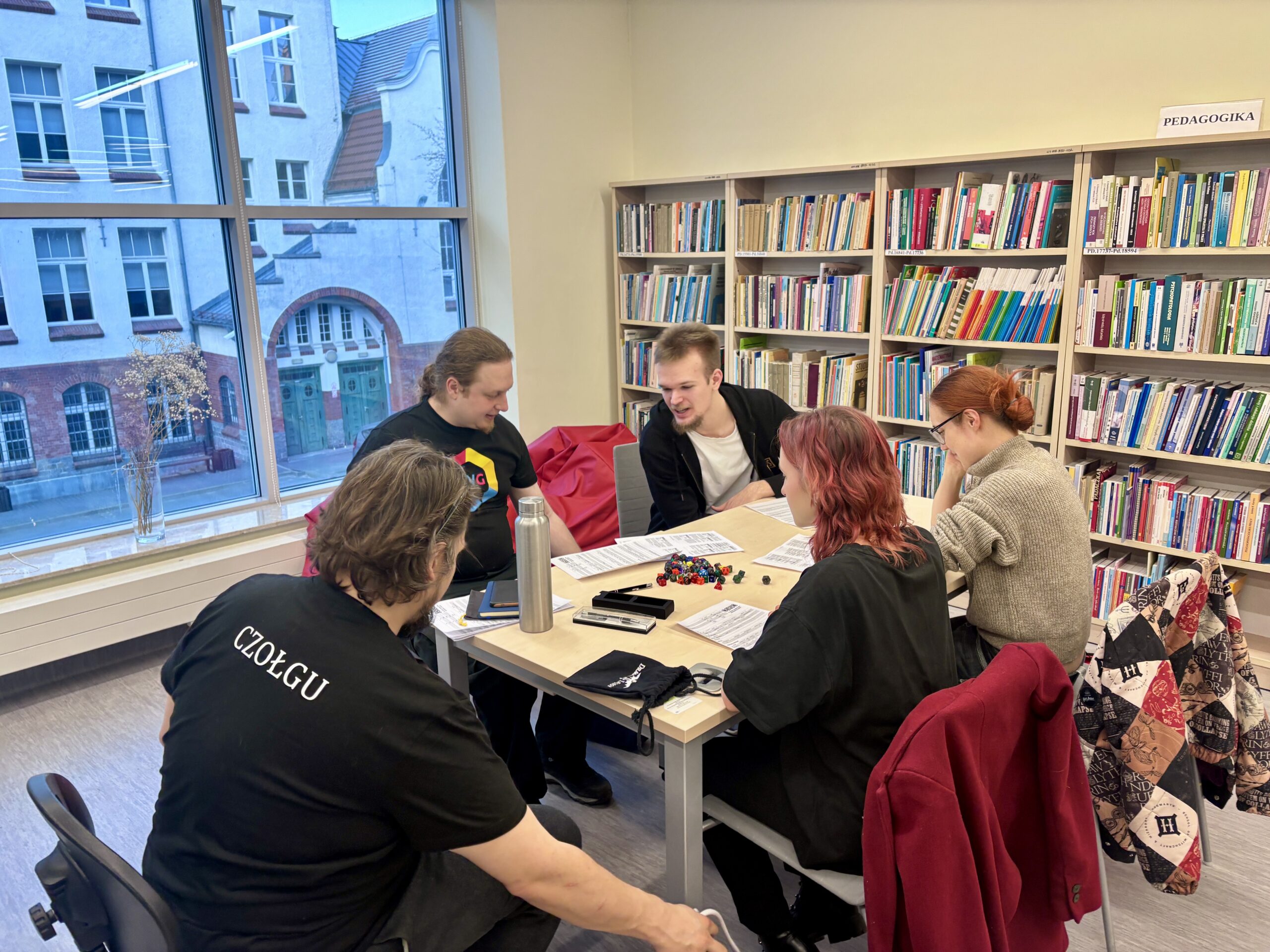 Pięć osób siedzących przy stole w sali bibliotecznej, grających w grę planszową.