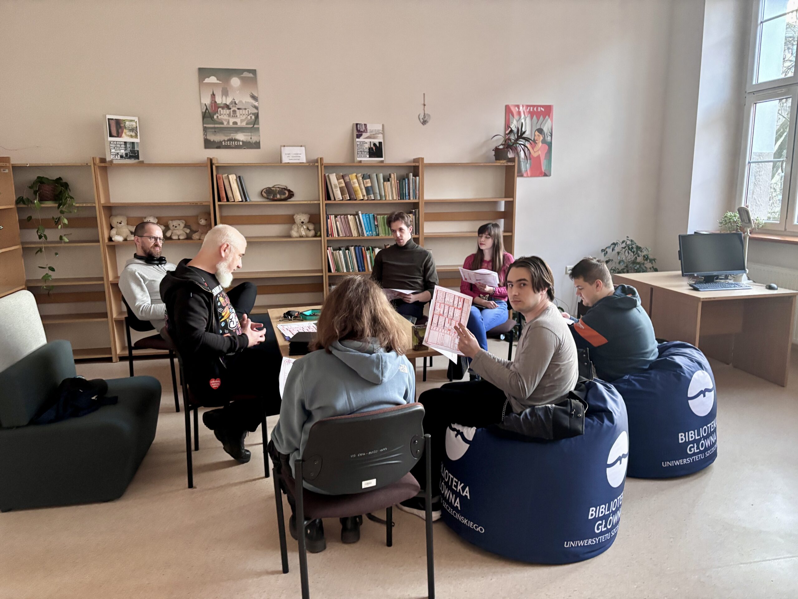 Grupa osób w sali bibliotecznej, siedząca na krzesłach i fotelach.. Część gra w grę planszową, część obserwuje tę grę.