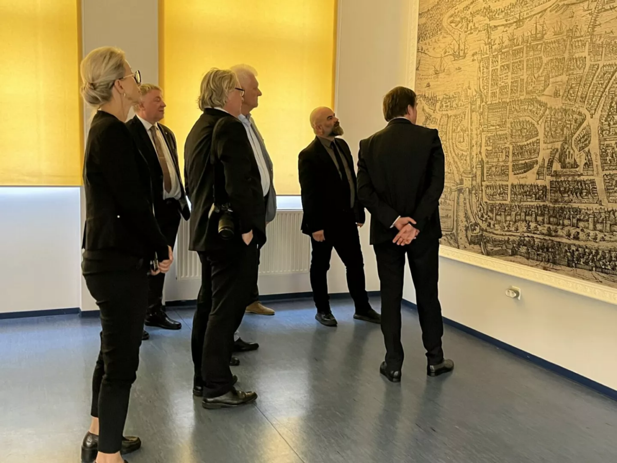 Po lewej sześcioro ludzi (bokiem do kadru), po prawej duża stara mapa, na którą patrzą.