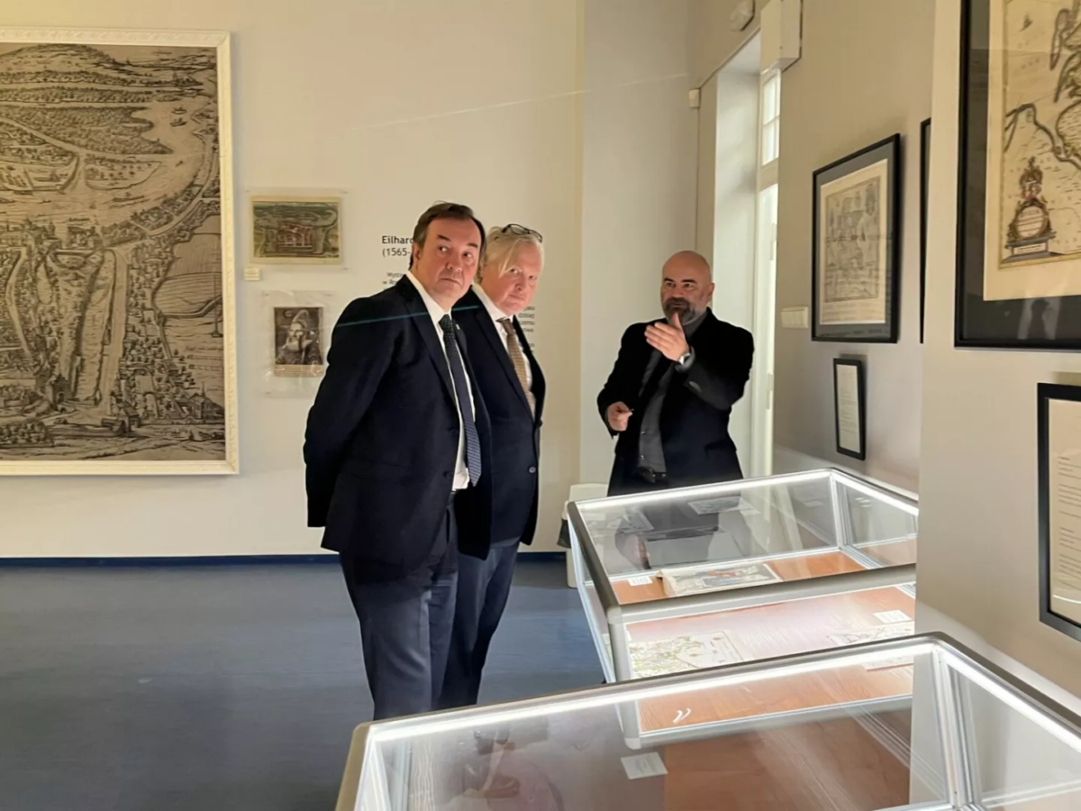 Na środku troje mężczyzn na wystawie Biblioteki Głównej Uniwersytetu Szczecińskiego. Po prawej przeszklone gabloty. Na ścianach stare mapy.