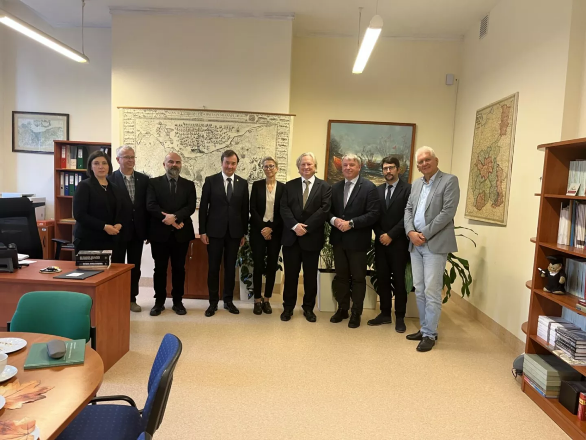 Dziewięcioro ludzi (w tym dwie kobiety) pozuje do zdjęcia, stojąc w jednym rzędzie. W tle na ścianach obrazy i stare mapy. Po lewej stół i biurko, po prawej regał z książkami.