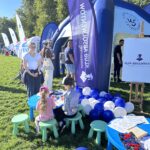 Po lewej stronie sztaluga z białą planszą z fioletowym logiem Klubu Absolwentów Uniwersytetu Szczecińskiego. Przy niej leżące na trawie granatowe i białe balony oraz stojące małe stoliki, przy których dzieci kolorują kolorowanki. W tle ludzie i granatowo-biały, półokrągły, dmuchany namiot z logami Uniwersytetu Szczecińskiego.