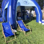 Stojący na trawie granatowo-biały, półokrągły, dmuchany namiot z logami Uniwersytetu Szczecińskiego. Pod namiotem ludzie. Przed nim dwa granatowe leżaki z okrągłym białym logiem US. W tle drzewa.