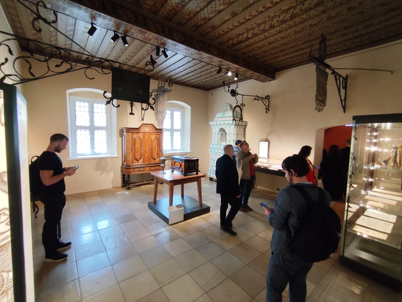 Wnętrze muzealnej sali z zabytkowymi meblami i kilkoma zwiedzającymi. Pomieszczenie ma jasne, kremowe ściany i dwa duże okna z kratowanymi szybami po lewej stronie. Na środku stoi drewniany stół ekspozycyjny na podwyższonej podstawie, a na nim ciemny, skrzyniowy lub szafkowy eksponat. Za stołem, pod oknami, znajduje się duża, ozdobna drewniana szafa o ciepłym, brązowym kolorze i dekoracyjnych frontach. Po prawej stronie, przy ścianie, stoi biały, masywny piec kaflowy z dekoracyjną górą. Wzdłuż prawej ściany ciągnie się niska, jasna gablotka z podświetlonymi dokumentami lub drobnymi eksponatami, a dalej widoczna jest przejściowa wnęka prowadząca do innego pomieszczenia. Na skrajnym prawym brzegu kadru widać wysoką, szklaną gablotę z podświetlonymi przedmiotami w środku. Sufit jest drewniany, z ciemnymi belkami i ozdobnymi malowaniami lub wzorami, a pod nim zamontowane są szyny oświetleniowe z reflektorami skierowanymi na eksponaty. Przy ścianach zawieszone są czarne, kute, ozdobne wsporniki lub elementy wystroju, częściowo z wiszącymi fragmentami tkaniny lub chorągwi. W sali jest kilka osób: po lewej mężczyzna w ciemnej koszulce, przy oknach i pośrodku dwie osoby rozmawiające, a bliżej prawej krawędzi stoi mężczyzna z plecakiem, trzymający telefon. Podłoga z dużych, jasnych płytek tworzy równą, gładką powierzchnię, a całość przestrzeni sprawia wrażenie spokojnej, muzealnej ekspozycji historycznej.