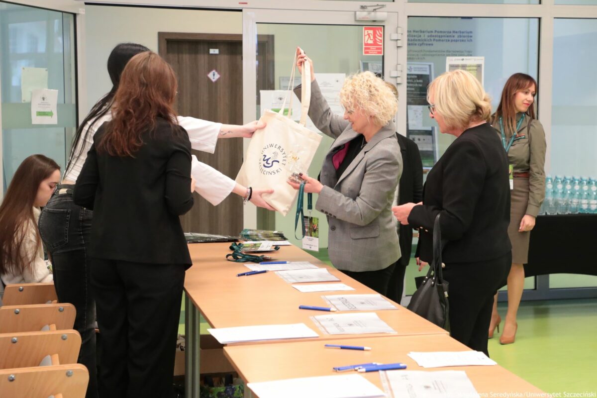 Scena rejestracji na konferencję lub wydarzenie na uczelni. Przy długim stole z jasnego drewna stoją osoby o jasnej i średniej karnacji, ubrane elegancko – głównie w marynarki w odcieniach szarości i czerni. Na pierwszym planie kobieta z kręconymi blond włosami w szarej marynarce odbiera płócienną torbę w kolorze ecru. Na torbie widoczne jest okrągłe logo i napis „Uniwersytet Szczeciński”. Obok niej stoi druga kobieta w czarnym żakiecie, trzymająca ciemną torebkę na ramieniu i patrząca w stronę stołu. Po przeciwnej stronie stołu stoją młodsze osoby, podające torbę i identyfikatory na smyczach. Na blacie stołu rozłożone są kartki formatu A4 w przezroczystych koszulkach, długopisy w niebieskim kolorze oraz kilka identyfikatorów na zielonych smyczach. W tle, po prawej stronie, stoi kobieta w szarej sukience służbowej z identyfikatorem na szyi, a za nią ustawione są rzędy butelek z wodą na czarnym stole. W głębi znajdują się szklane ściany i drzwi z naklejkami informacyjnymi oraz znak przeciwpożarowy.