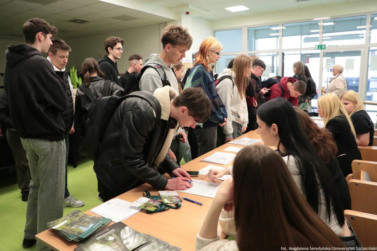 Grupa młodych osób w kolejce przy stole rejestracyjnym. W środku kadru kilka osób stoi wzdłuż długiego stołu, część pochylona nad dokumentami, niektórzy piszą lub podpisują papiery. Przy stole siedzi kilka kobiet, podają kartki i wskazują miejsca do wypełnienia, na blacie leżą długopisy, broszury i identyfikatory na smyczach. Większość osób ma plecaki i ubrania w stylu codziennym: bluzy, kurtki, dżinsy. W tle widać jasne, przeszklone drzwi i okna prowadzące na korytarzu budynku, a dalej pojedyncze osoby przechodzące obok.
