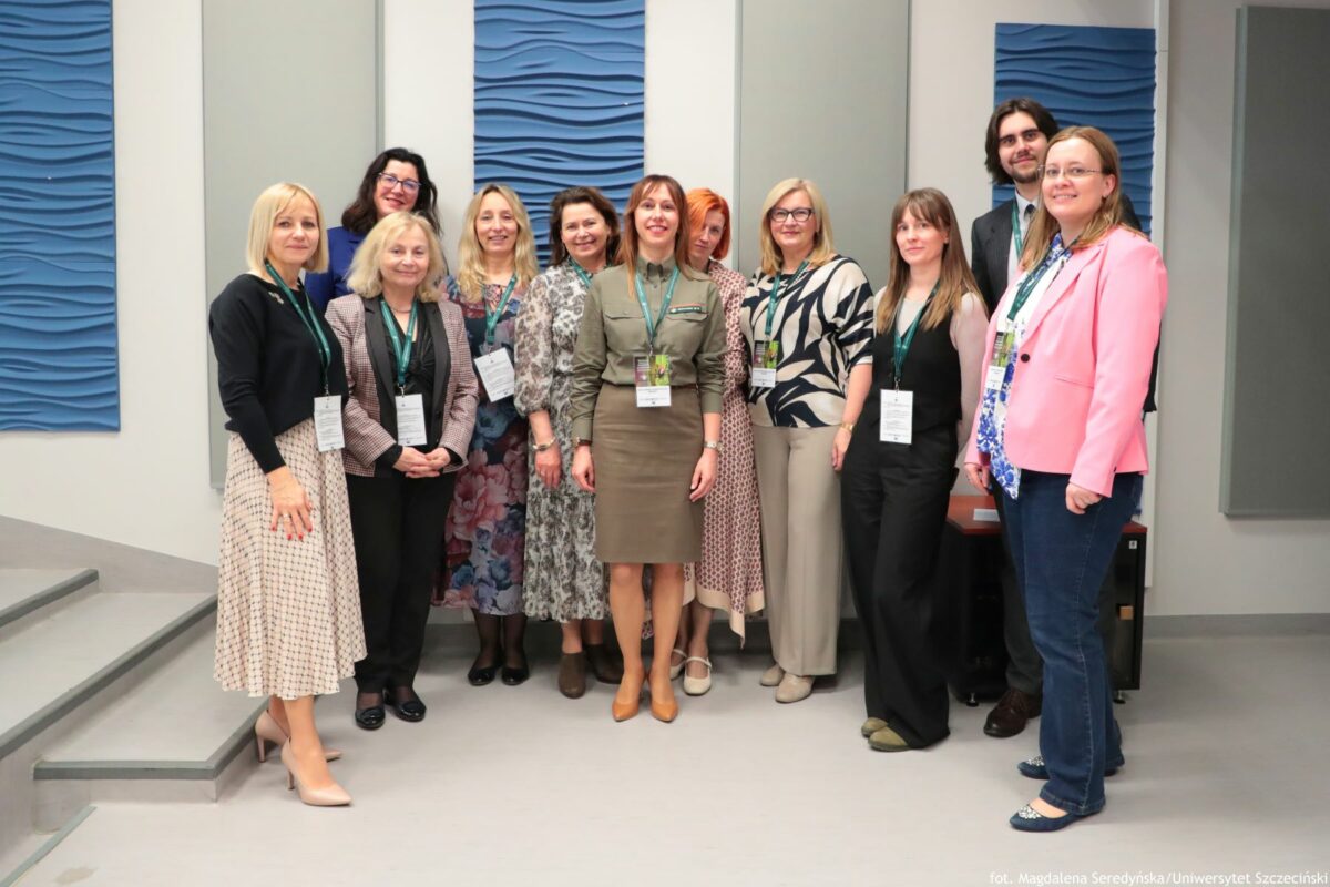 Grupa jedenastu osób stojących obok siebie w pomieszczeniu. Wszyscy stoją przodem do aparatu i uśmiechają się. Dziesięć osób to kobiety, jedna osoba to mężczyzna stojący z tyłu po prawej stronie. Skóra wszystkich ma jasny odcień. Osoby mają na szyjach zielone smycze z identyfikatorami, co sugeruje konferencję lub spotkanie zawodowe. Od lewej kobieta w czarnym sweterku i jasnej, wzorzystej, za kolano spódnicy, w beżowych butach na obcasie. Obok stoi niższa kobieta o jasnych, półdługich włosach, w kratkowanej marynarce, czarnych spodniach i czarnych butach. Dalej widać kobietę w długiej, kwiecistej sukience w odcieniach różu i fioletu z czarnymi rajstopami i czarnymi butami. Kolejna kobieta ma na sobie długą, jasnoszarą, wzorzystą sukienkę i brązowe buty. Na środku stoi kobieta w oliwkowej sukience koszulowej do kolan z długim rękawem i paskiem, w jasnobrązowych butach na obcasie. Po jej prawej stronie jest kobieta o rudych włosach, w jasnej sukience z drobnym wzorem i jasnych butach. Obok niej stoi kobieta w okularach, w luźnej, beżowo-granatowej bluzce z abstrakcyjnym wzorem, jasnych, szerokich spodniach i jasnych butach. Dalej po prawej widać kobietę w błękitnej koszulce pod czarnym kombinezonem na ramiączkach, w zielonych butach. Obok niej stoi mężczyzna w ciemnym garniturze, białej koszuli i krawacie, częściowo zasłonięty, widoczne są głowa, ramiona i fragment nóg. Na skraju prawej strony znajduje się kobieta w różowej marynarce, granatowych dżinsach, niebiesko-białej koszuli i balerinach w ciemnym kolorze.