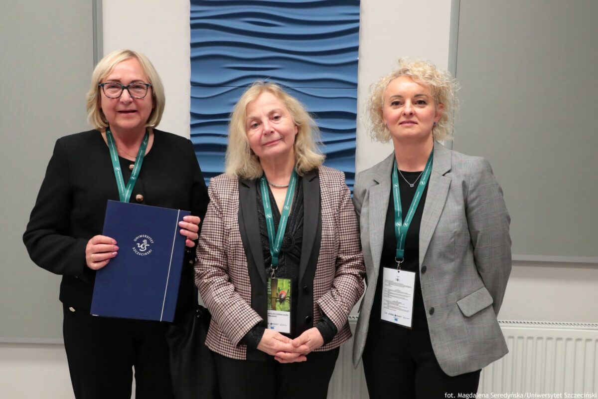 Trzy kobiety stoją obok siebie na tle jasnej ściany z dekoracyjnym, niebieskim panelem o pofalowanej fakturze pośrodku. Wszystkie mają jasną karnację i noszą na szyi zielone smycze konferencyjne z identyfikatorami. Kobieta po lewej ma krótkie, jasnoblond włosy, okulary i czarną, elegancką sukienkę lub garsonkę zapinaną na złote guziki. W dłoniach trzyma granatową teczkę lub segregator z białym napisem „UNIWERSYTET SZCZECIŃSKI” i logo uczelni. Kobieta w środku ma dłuższe, jasne włosy do ramion, lekko falowane. Ubrana jest w czarną bluzkę i marynarkę w drobną kratkę, z czarnymi wykończeniami przy klapach. Na identyfikatorze zawieszonym na smyczy widać zdjęcie z zielonym tłem i postacią w czerwonym stroju. Kobieta po prawej ma krótkie, mocno kręcone blond włosy. Ubrana jest w szarą marynarkę, czarną bluzkę i ciemne spodnie. Na identyfikatorze przewieszonym na smyczy widoczny jest tekst w kilku wierszach. W tle po bokach znajdują się gładkie panele ścienne w jasnoszarym kolorze oraz fragment białego grzejnika na dole po prawej stronie. W prawym dolnym rogu zdjęcia drobnym drukiem znajduje się podpis: „fot. Magdalena Seredyńska/Uniwersytet Szczeciński”.