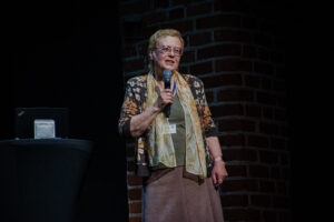 Czarne tło. Po prawej widać fragment ściany z czerwonej cegły. Na scenie kobieta w blond krótkich włosach i okularach trzymająca mikrofon. Po lewej stronie wysoki stolik z czarnym skirtingiem, na którym stoi laptop.