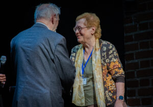 Czarne tło. Po prawej widać fragment ściany z czerwonej cegły. Na scenie kobieta w blond krótkich włosach i okularach (ujęcie do pasa). Po lewej stronie tyłem do kadru mężczyzna w szarej marynarce ściska dłoń kobiety.