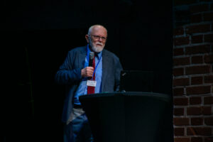 Czarne tło. Po prawej widać fragment ściany z czerwonej cegły. Na scenie mężczyzna trzymający mikrofon. Przed nim wysoki stolik z czarnym skirtingiem, na którym stoi laptop.