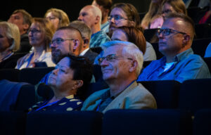 Ujęcie bokiem. Widownia na ciemnych fotelach. Kilkanaście osób. Na pierwszym planie uśmiechnięty starszy mężczyzna w okularach.