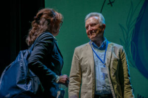 Zielone tło. Po prawej stronie uśmiechnięty mężczyzna w jasnej marynarce i siwych włosach (ujęcie do pasa). Po lewej stronie bokiem do kadru kobieta z plecakiem, która – trzymając wodę w butelce – rozmawia z mężczyzną.