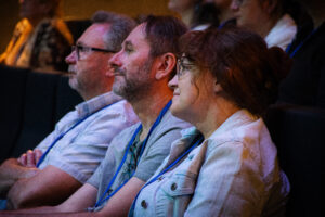 Ujęcie bokiem. Widownia na ciemnych fotelach. Trzy osoby. Na pierwszym planie uśmiechnięta kobieta w okularach we włosach spiętych w kok.
