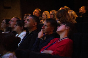 Ujęcie bokiem. Widownia na ciemnych fotelach. Kilkanaście osób. Na pierwszym planie kobieta w czerwonej bluzce z głową opartą o oparcie i mężczyzna podpierający dłonią podbródek.