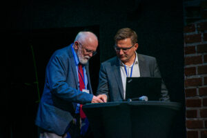 Czarne tło. Po prawej widać fragment ściany z czerwonej cegły. Na scenie dwóch mężczyzn. Przed nimi wysoki stolik z czarnym skirtingiem, na którym stoi laptop i leży mikrofon.