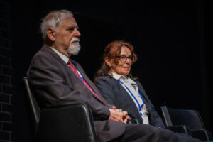 Czarne tło. Po lewej widać fragment ściany z czerwonej cegły. Na scenie siedzą na krzesłach mężczyzna i kobieta. Ujęcie bokiem.