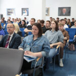 Sala wykładowa wypełniona siedzącymi osobami. W kilku rzędach ustawione są krzesła konferencyjne z niebieskimi i szarymi tapicerowanymi siedziskami oraz jasnymi podłokietnikami. Najbliżej obiektywu siedzi mężczyzna w garniturze, z okularami i krawatem w bordowym kolorze; obok niego z przodu siedzi młoda kobieta w jasnoniebieskiej, dżinsowej kurtce, z ciemnymi włosami do ramion, trzyma na kolanach teczki i przezroczystą butelkę wody. Za nimi widoczni są głównie młodzi dorośli, wiele osób ma na sobie codzienne ubrania, takie jak T‑shirty, bluzy, koszule i dżinsy; większość patrzy przed siebie, kilka osób spogląda w dół na telefony lub notatki. Ściany są białe, na nich wiszą kilka obrazów lub fotografii w ramach. Podłoga jest pokryta niebieską wykładziną. Wrażenie jest takie, jak podczas wykładu lub prezentacji na uczelni.