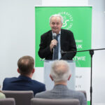 Starszy mężczyzna o jasnych, przerzedzonych włosach stoi za przeźroczystą mównicą i przemawia do publiczności, trzymając mikrofon w prawej dłoni. Ma jasną karnację. Ubrany jest w czarną marynarkę, jasnoniebieską koszulę i ciemny krawat, w brustaszy widać ciemną poszetkę. Przed nim siedzą na krzesłach trzy osoby odwrócone tyłem do aparatu: kobieta z dłuższymi brązowymi włosami po lewej oraz dwóch mężczyzn o siwych i jasnych włosach pośrodku i po prawej, wszyscy w formalnych ubraniach. Za mównicą znajduje się zielona tablica lub baner z białym logo i napisem „Uniwersytet Szczeciński” oraz kilkoma innymi białymi słowami w języku polskim. Po prawej stronie z przodu zamocowany jest statyw z długim mikrofonem skierowanym w stronę mówiącego. Tło stanowi jasna, gładka ściana we wnętrzu sali konferencyjnej lub wykładowej.