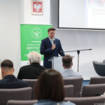 Sala konferencyjna z mówiącym przy mównicy mężczyzną w garniturze. Mężczyzna stoi za przezroczystą mównicą, trzyma mikrofon i przemawia do siedzącej publiczności. Za nim po lewej stronie znajduje się zielony baner z logo „Uniwersytet Szczeciński” oraz napisem „Instytut o Polityce i Bezpieczeństwie” (środkowa część tekstu częściowo zasłonięta). Nad banerem wisi godło Polski z białym orłem na czerwonym tle. Po prawej stronie wisi ekran projekcyjny, na którym widać fragment okładki publikacji z tytułem „Kronika Szczecina 2024” i dalszym, słabiej czytelnym tekstem poniżej. Przed mównicą siedzi kilka osób tyłem do aparatu, na szarych krzesłach konferencyjnych. W dolnym prawym rogu widać drobny biały napis „fot. Magdalena Seredyńska/Uniwersytet Szczeciński”.