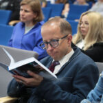 Mężczyzna w okularach czytający książkę w sali wykładowej. Ma krótkie, przerzedzone włosy w kolorze jasnobrązowym i jasną karnację. Nosi ciemnoniebieską marynarkę z miękkiego materiału, jasną koszulę i bordowo-granatowy krawat w paski. Trzyma otwartą, dość grubą książkę w miękkiej oprawie, nachyloną blisko twarzy; strony są białe, na okładce widać fragment kolorowej fotografii, ale bez czytelnego tytułu. Siedzi na jasnoszarym, miękkim fotelu z jasnym podłokietnikiem. W tle znajdują się inne osoby siedzące w rzędach na niebieskich fotelach. Bezpośrednio za mężczyzną siedzi kobieta o jasnej cerze i krótkich, jasnobrązowych włosach, ubrana w niebieską koszulę. Dalej z prawej strony widoczna jest kobieta o bardzo jasnych, długich, prostych włosach, w okularach i czarnej marynarce. Tło jest lekko rozmyte, ale sugeruje wykład lub spotkanie w dużej sali z wieloma uczestnikami.