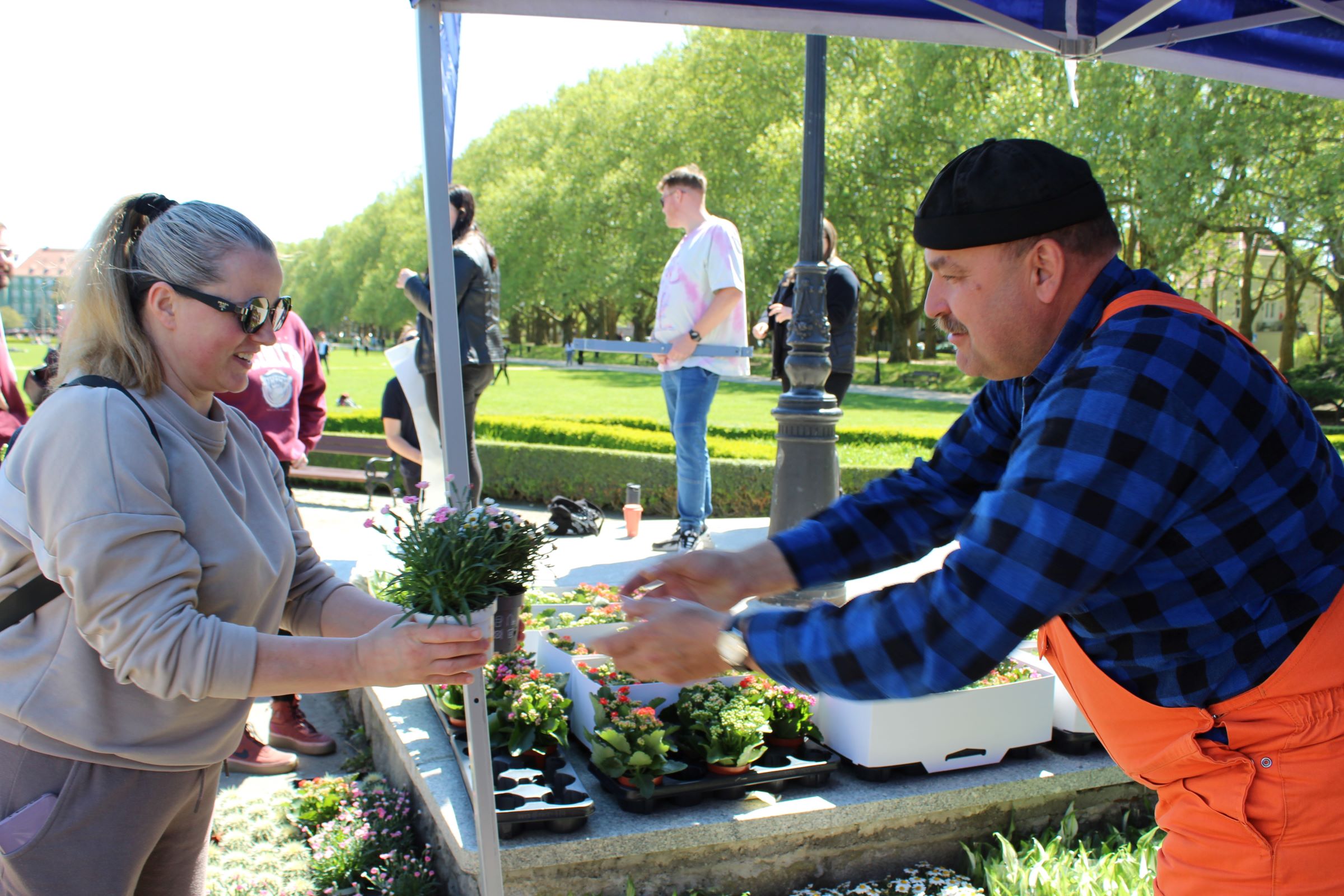 Stoisko z kwiatami na zewnątrz, pod niebiesko-białym daszkiem, z dwiema osobami w centrum kadru podczas przekazywania doniczki z rośliną. Po lewej stronie jest kobieta o jasnej karnacji i blond włosach związanych w kucyk; ma ciemne okulary przeciwsłoneczne i jasnoszarą bluzę. Trzyma w obu rękach małą doniczkę z zieloną rośliną i drobnymi fioletowymi kwiatami. Po prawej stronie jest mężczyzna o jasnej karnacji, w czarnym berecie, z wąsem, w niebiesko-czarnej koszuli w kratę i pomarańczowych ogrodniczkach; wyciąga ręce w stronę doniczki, jakby ją podawał lub odbierał. Na blacie przed nimi stoją tacki z małymi roślinami i kwiatami w różnych kolorach, a także białe skrzynki/pojemniki. W tle widać park z zielonym trawnikiem, żywopłotami i drzewami oraz kilka osób stojących lub przechodzących; obok jest czarna latarnia parkowa.